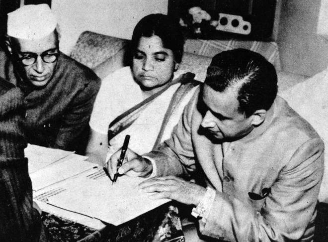 Durgabai Deshmukh seated between two men at a low table; the man on the right signs a document with a fountain pen while she watches.