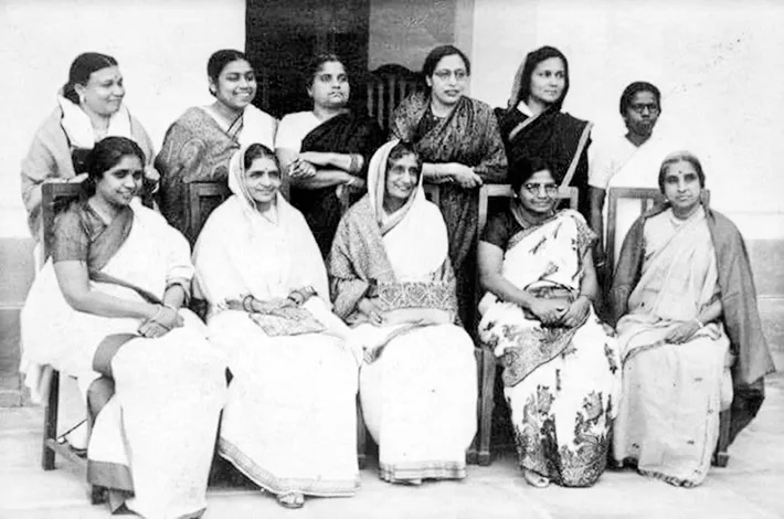 Group photograph of eleven women in saris, six seated on chairs in front and five standing behind, on a veranda.