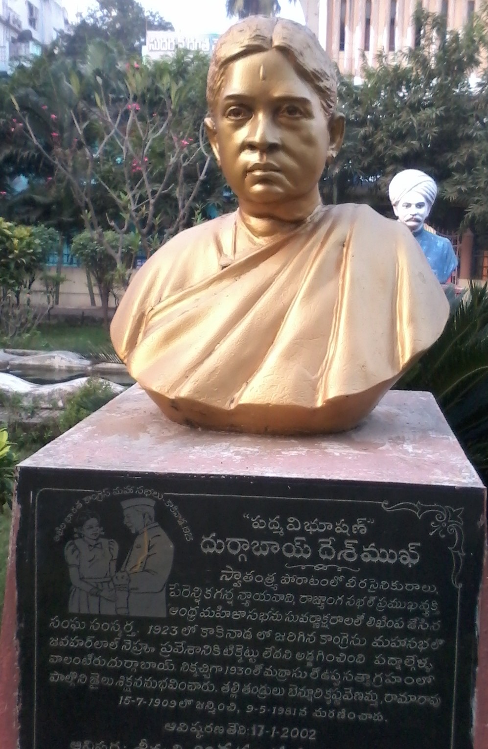 A gilded bronze bust of Durgabai Deshmukh on a stone plinth in a garden, with a Telugu inscription carved into the black plinth below.