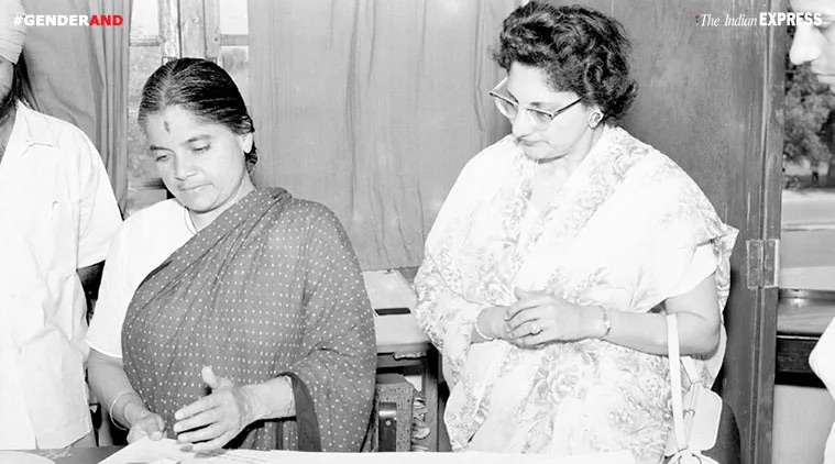 Durgabai Deshmukh, on the left in a dark patterned sari, standing at a table beside another woman in a light sari and spectacles, both looking down at papers.